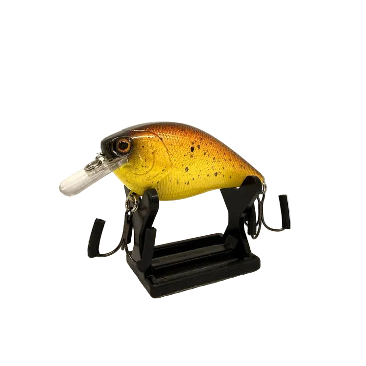 A fishing lure displayed on a black stand against a white textured background. The lure has a yellow and orange body with black speckles, a dark head, and a clear plastic lip.